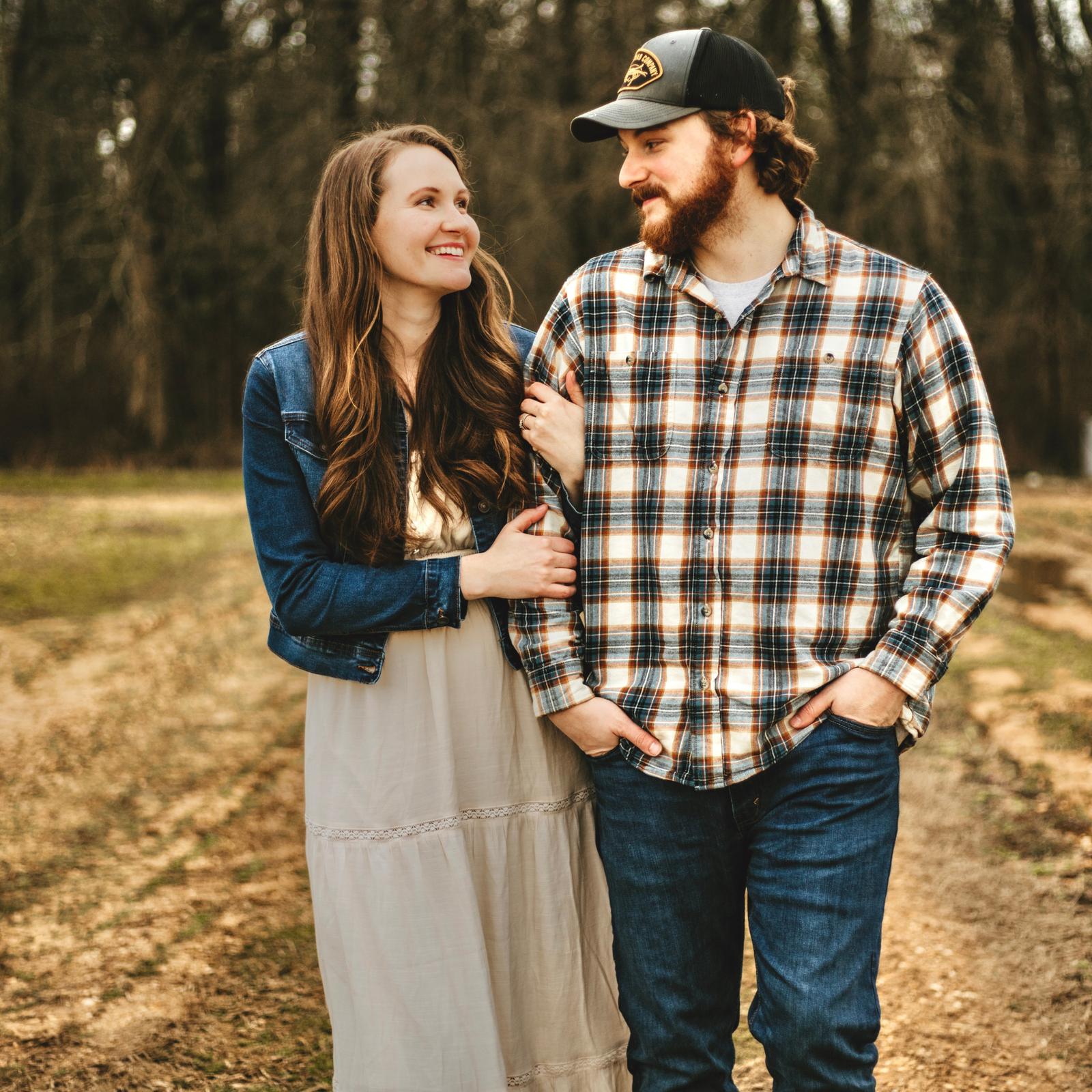 female in white dress and jean jacket in arm of man with flannel happily walking on a farm